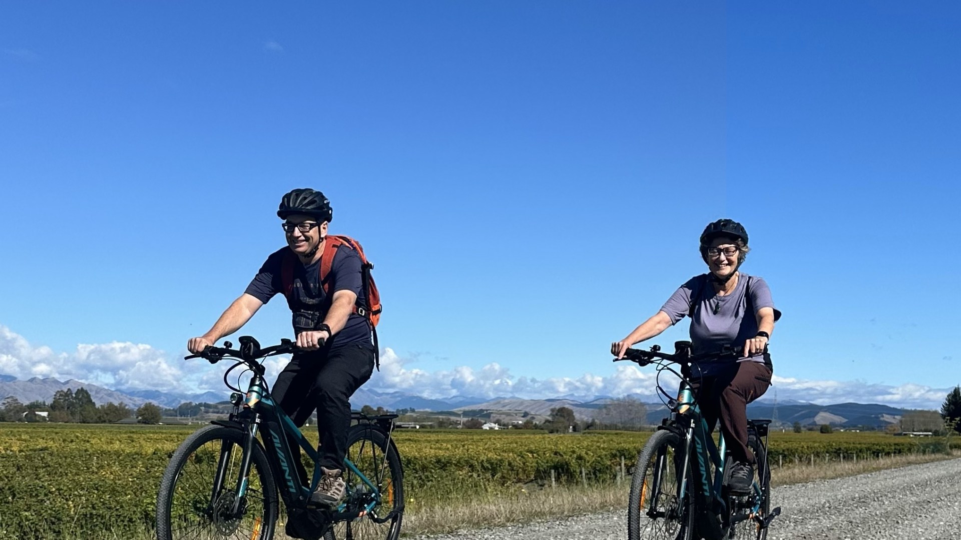 Two cyclists with helmets riding on a gravel path under a clear blue sky.