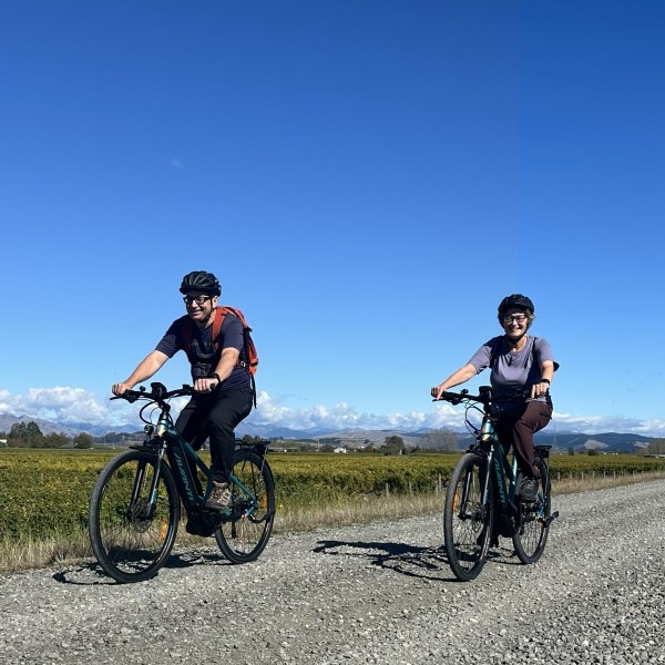 Two cyclists with helmets riding on a gravel path under a clear blue sky.