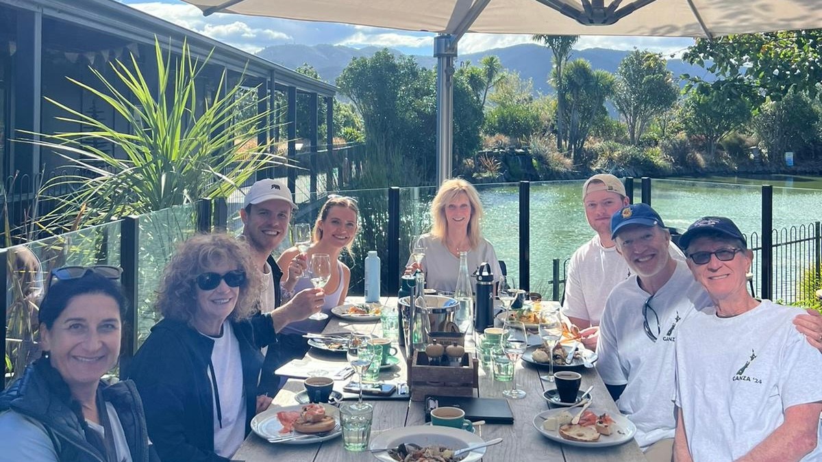 A group of people dining outside at a long table under a large umbrella with a pond and greenery in the background.