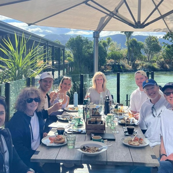 A group of people dining outside at a long table under a large umbrella with a pond and greenery in the background.