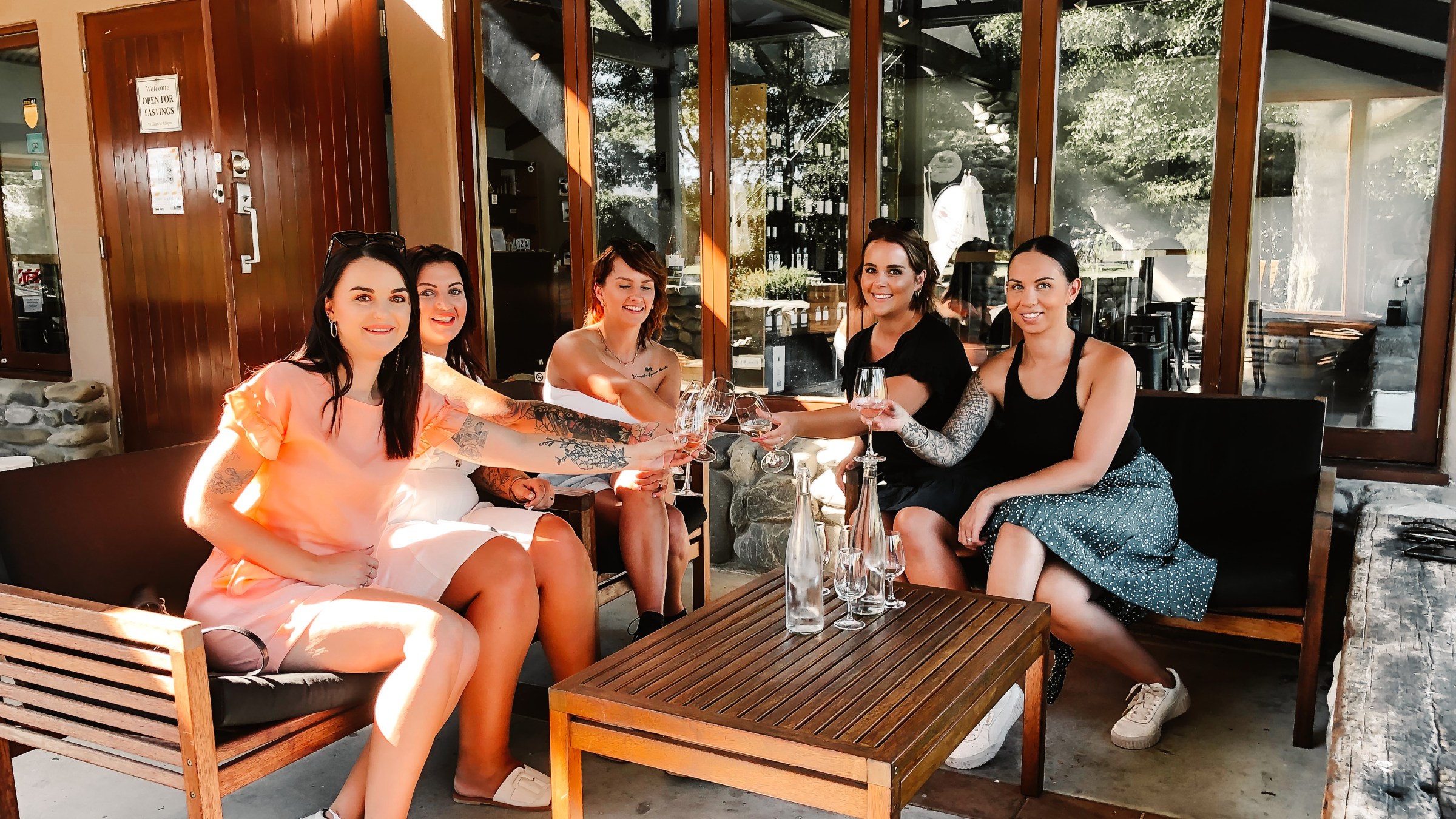 Five women sitting outside, toasting with glasses in a relaxed setting.
