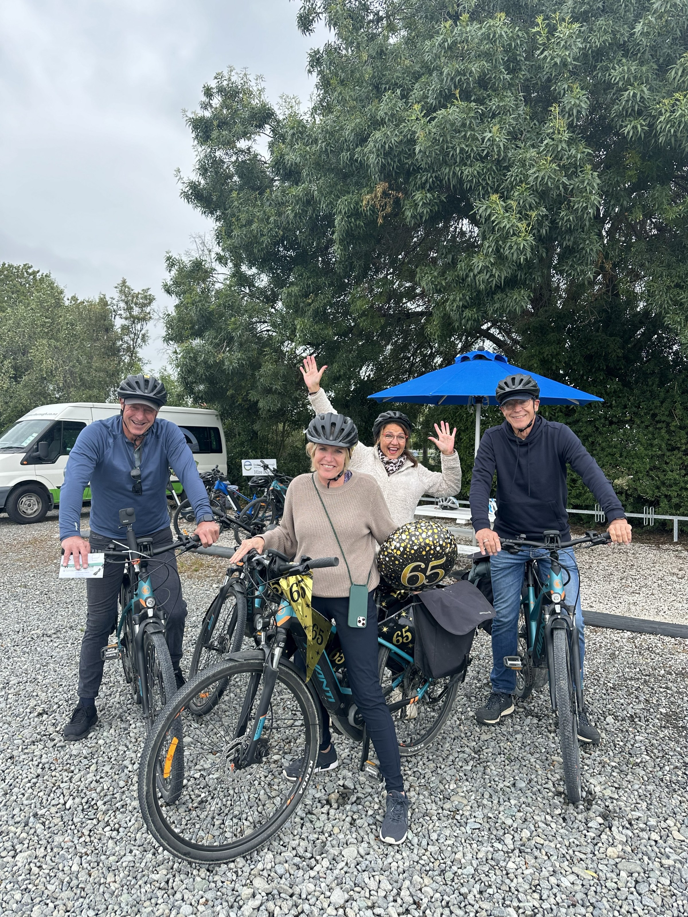 Four people with helmets posing with bikes and '65' balloons outdoors.