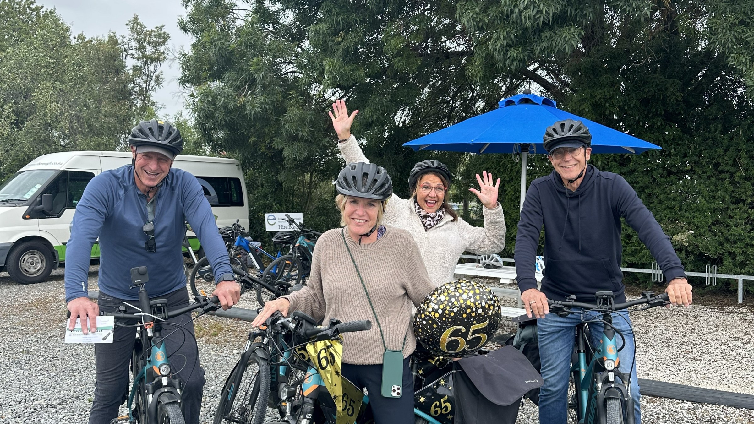 Four people with helmets posing with bikes and '65' balloons outdoors.