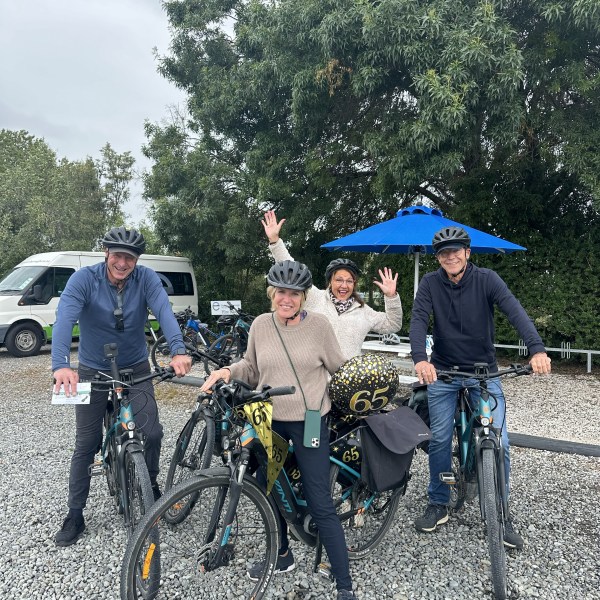 Four people with helmets posing with bikes and '65' balloons outdoors.