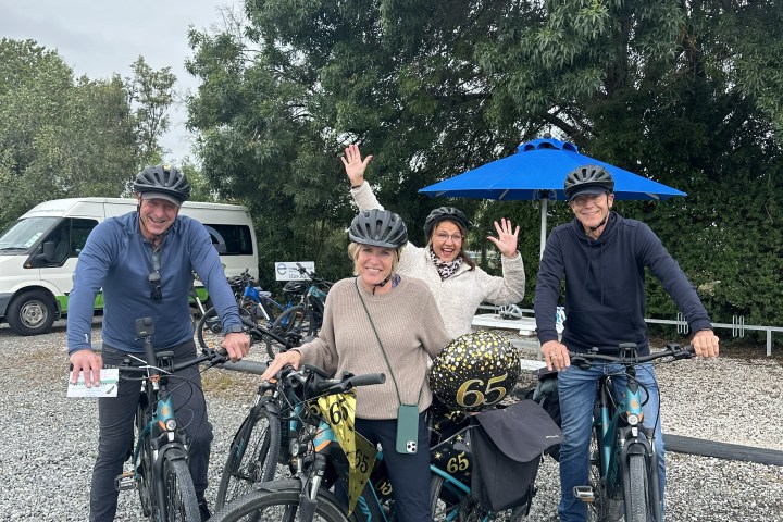 Four people with helmets posing with bikes and '65' balloons outdoors.