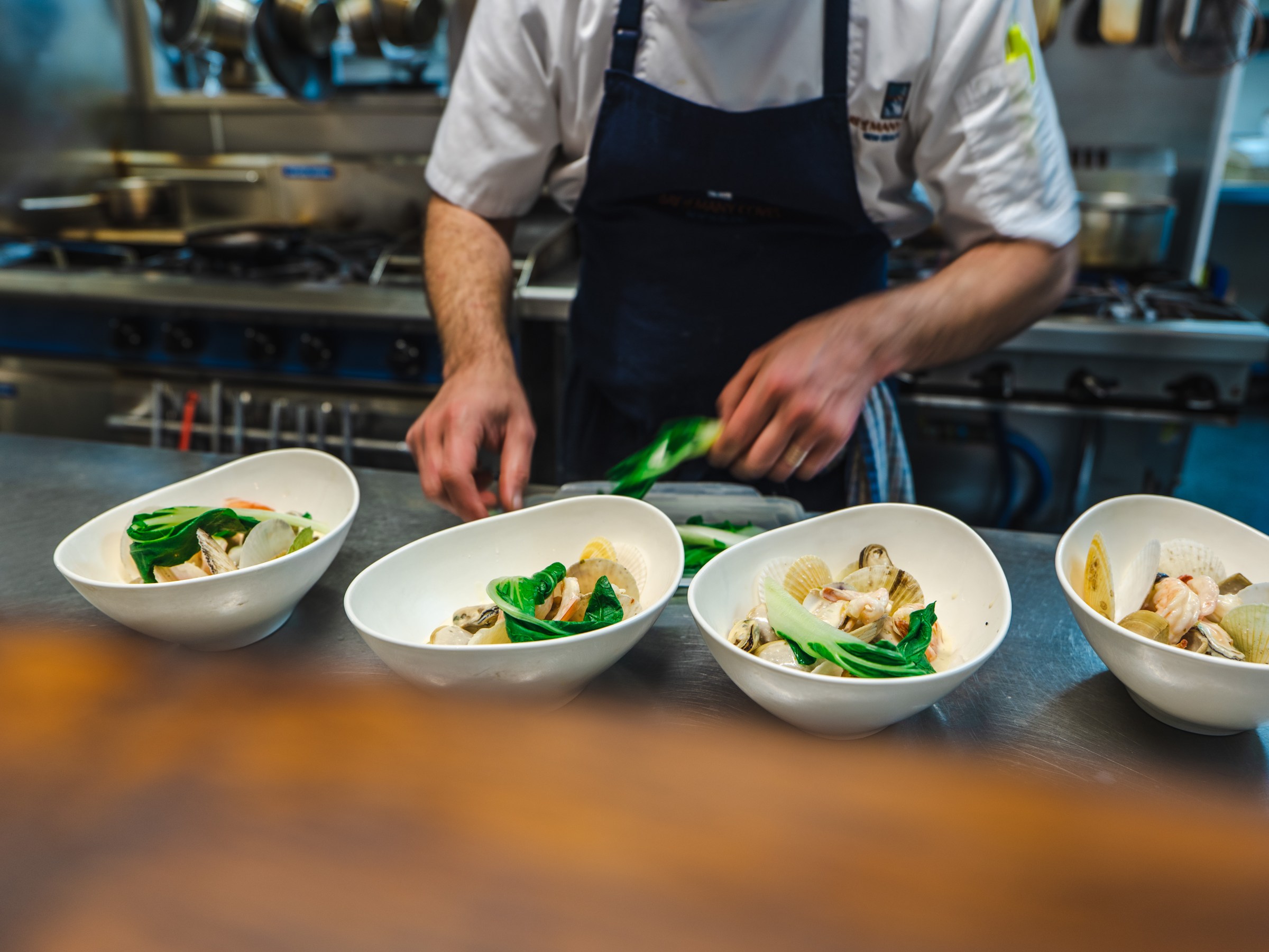 Chef plating seafood dish in white bowls on stainless steel counter in a kitchen.