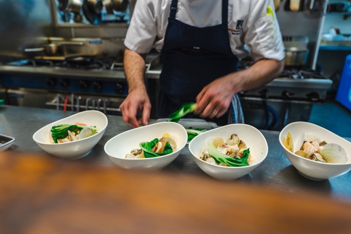 Chef plating seafood dish in white bowls on stainless steel counter in a kitchen.