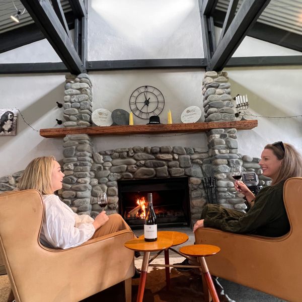 Two women sit in armchairs by a stone fireplace, drinking red wine in a cozy room.