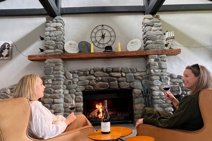 Two women sit in armchairs by a stone fireplace, drinking red wine in a cozy room.