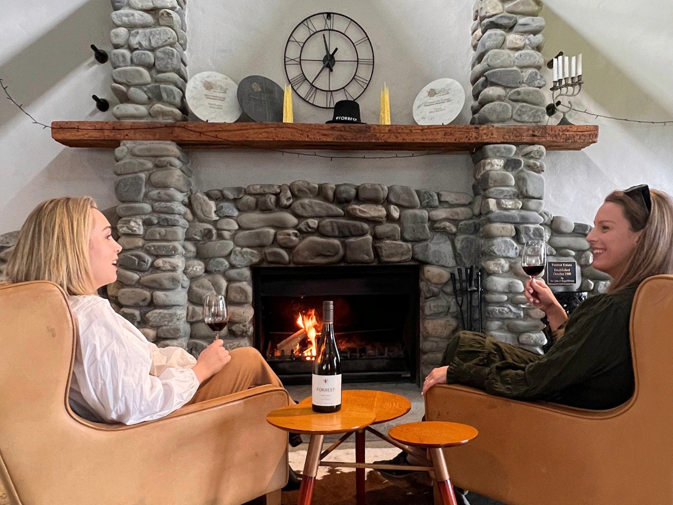 Two women sit by a stone fireplace, holding wine glasses and smiling.