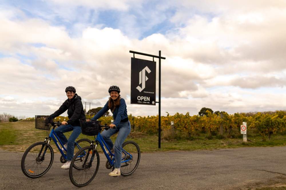 Two people biking past a vineyard with a sign that reads 'Open' under a cloudy sky.