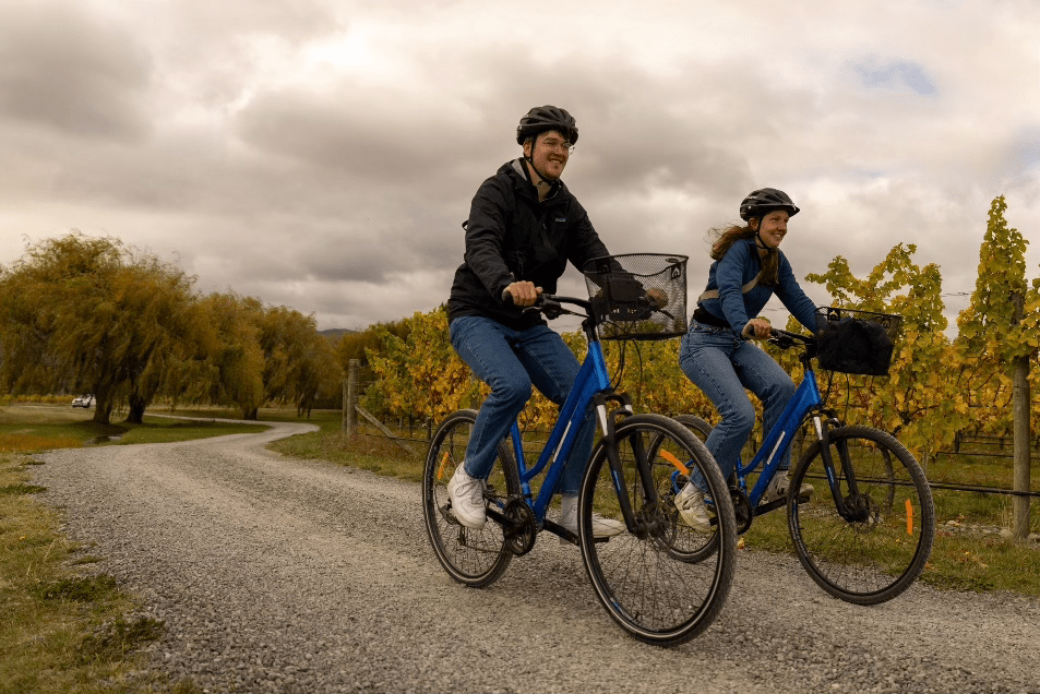 Two people cycling on a gravel path surrounded by trees and vineyards under a cloudy sky.