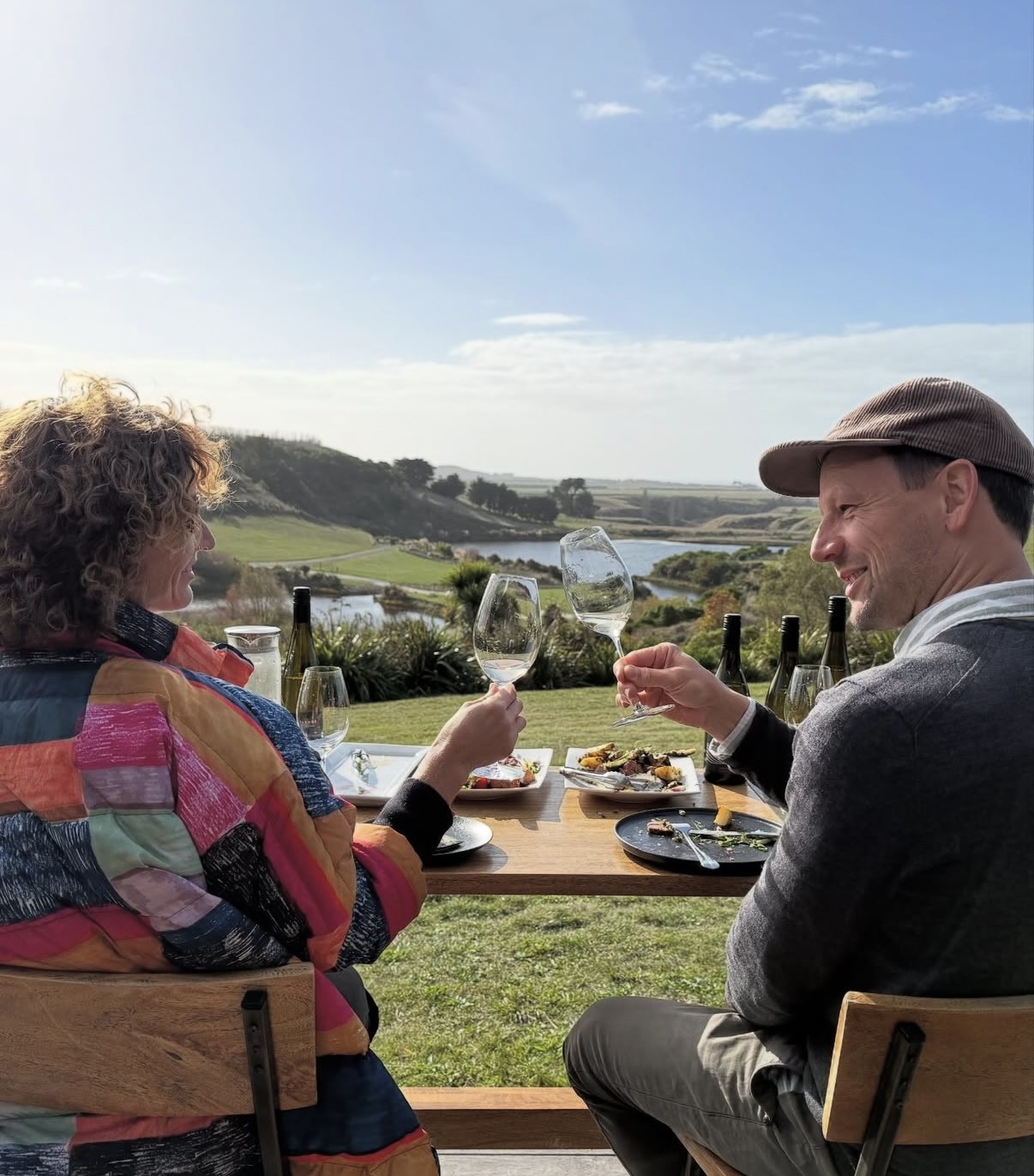 Two people toast with wine glasses at an outdoor table overlooking a scenic landscape.