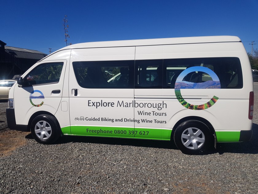 White van with 'Explore Marlborough Wine Tours' logo parked on gravel under a blue sky.