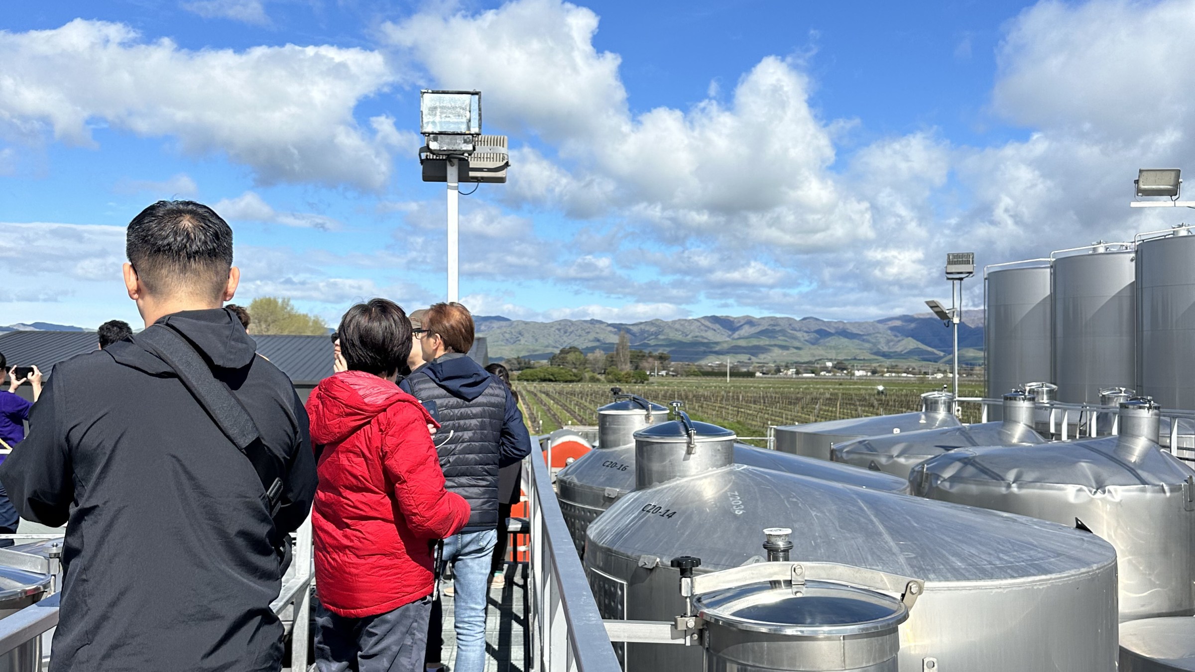 People on a walkway above industrial tanks with a clear blue sky and distant hills.