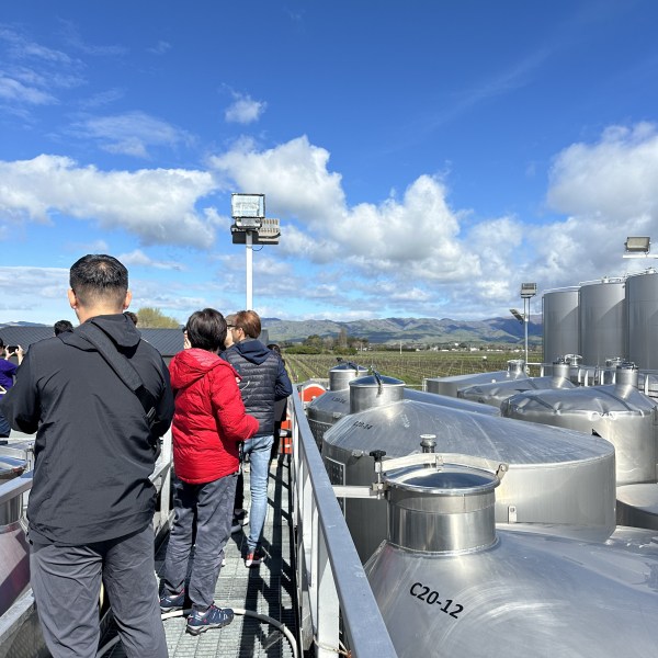 People on a walkway above industrial tanks with a clear blue sky and distant hills.
