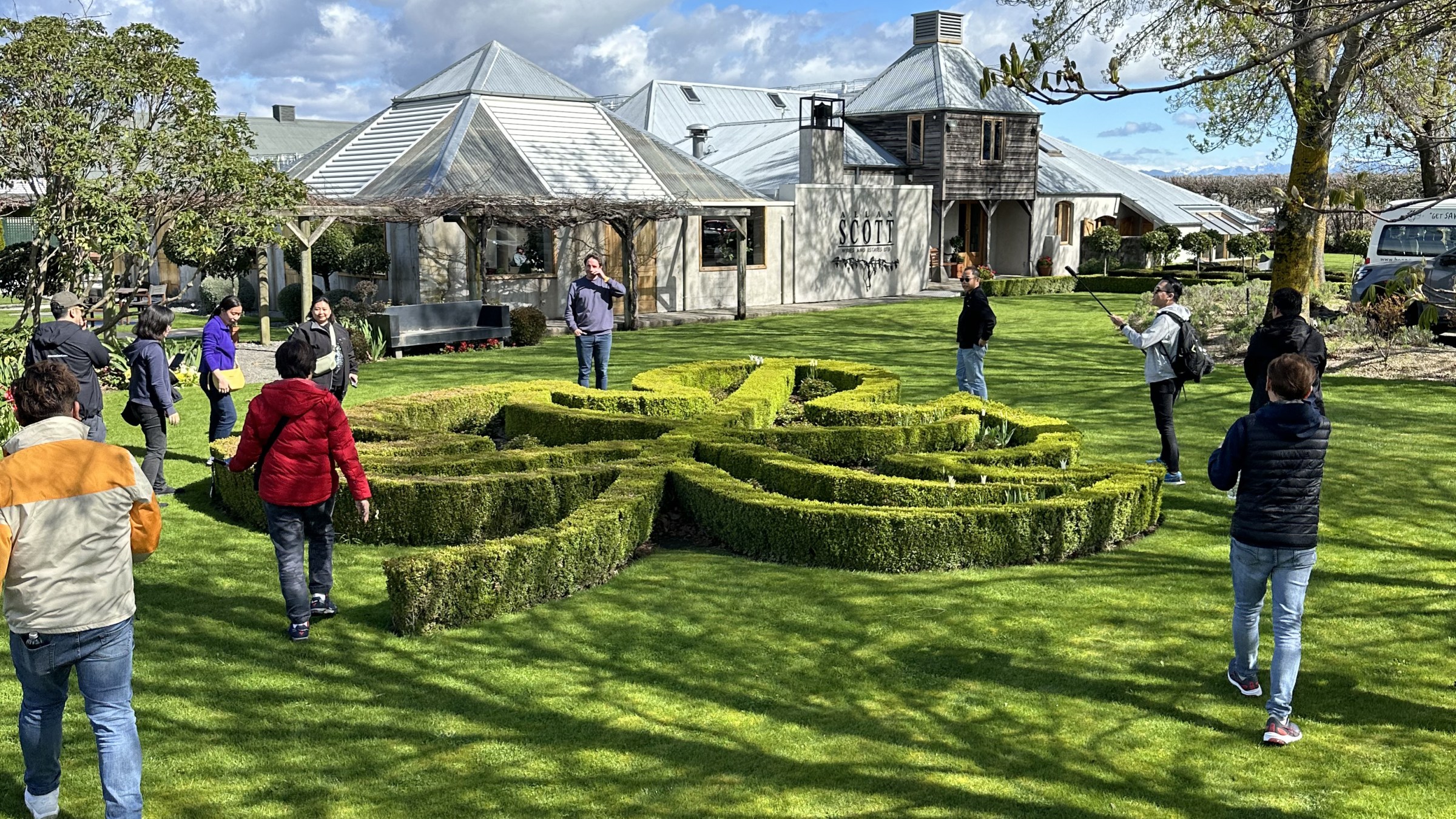 People walking on grass near a large building and decorative hedges under a blue sky.
