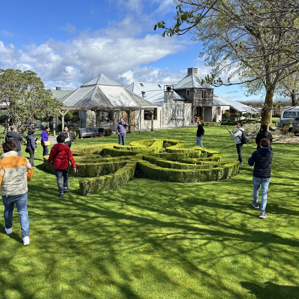 People walking on grass near a large building and decorative hedges under a blue sky.