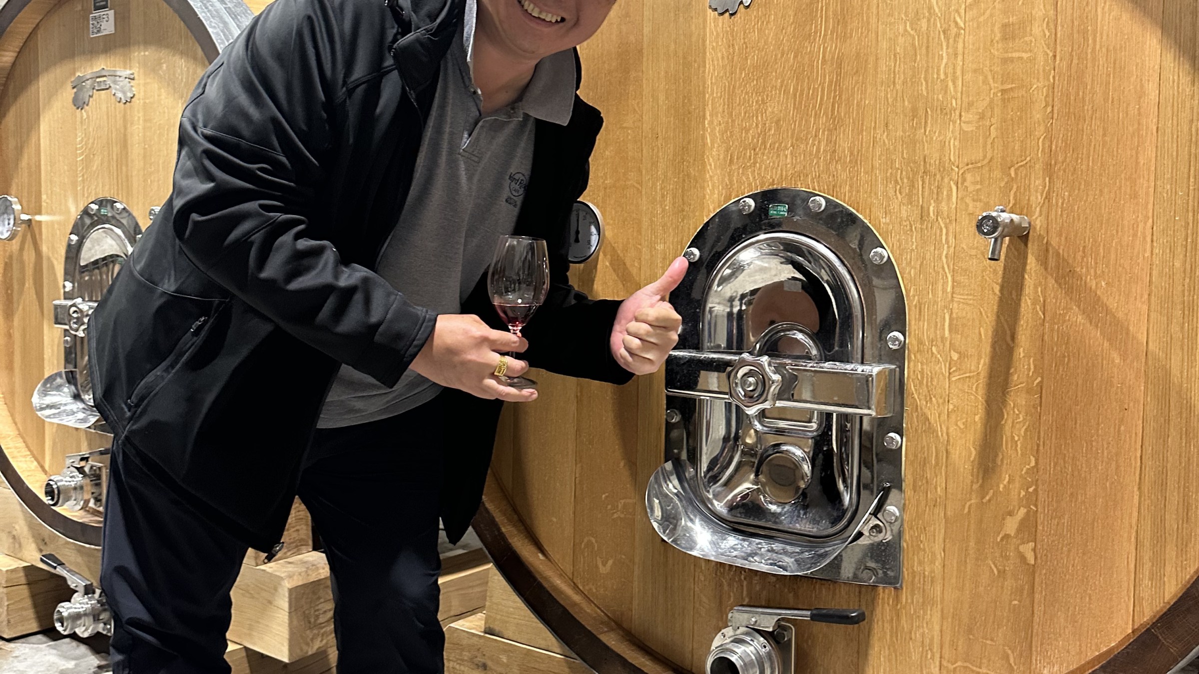 Person smiling with wine glass, standing by large wooden barrel in a cellar.