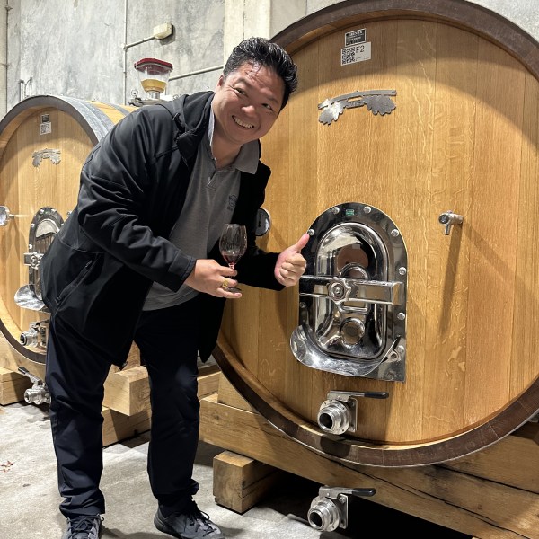Person smiling with wine glass, standing by large wooden barrel in a cellar.