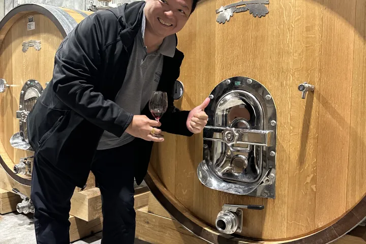 Person smiling with wine glass, standing by large wooden barrel in a cellar.