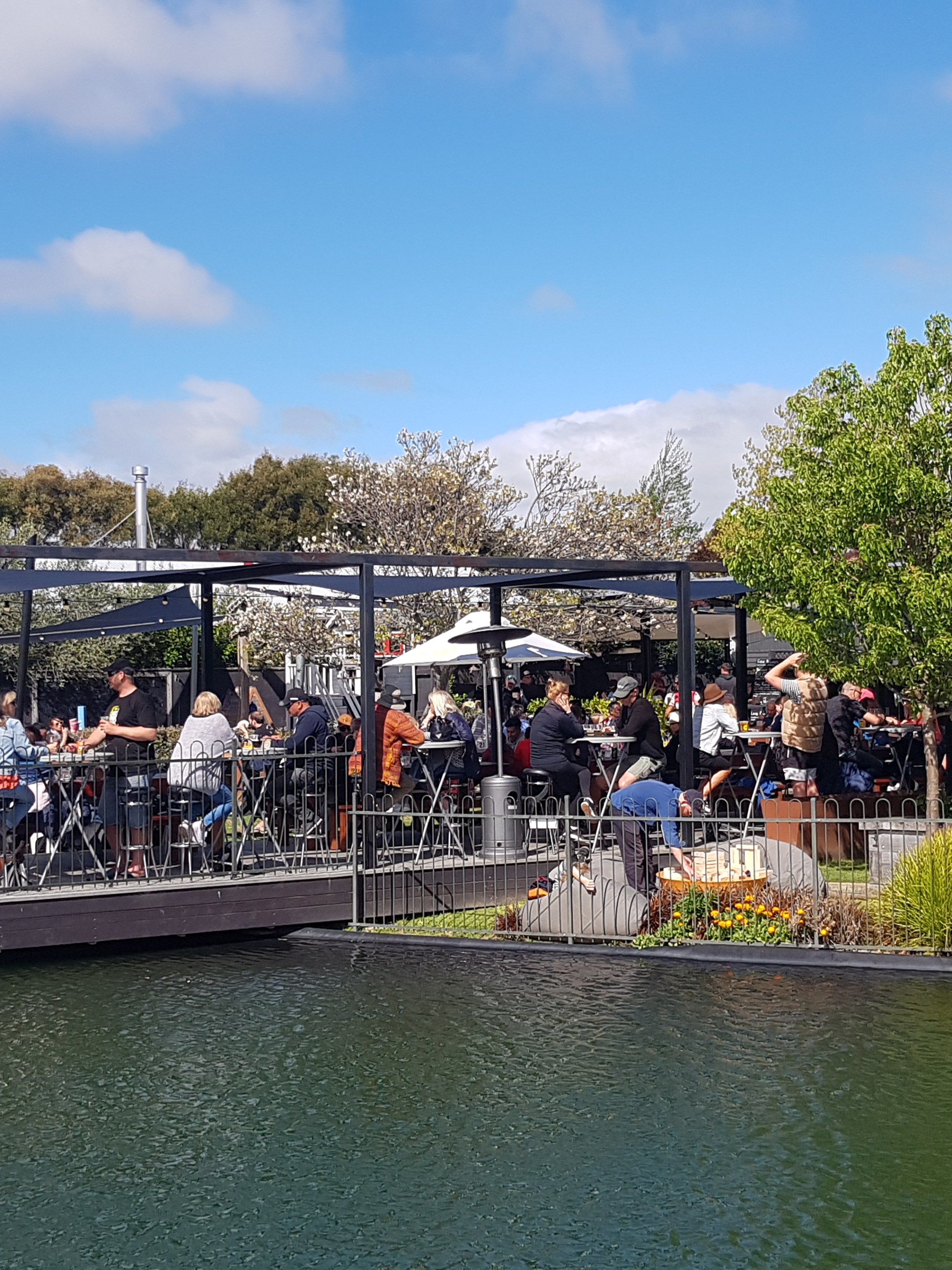 Outdoor dining area beside a pond with people seated under umbrellas and pergolas.