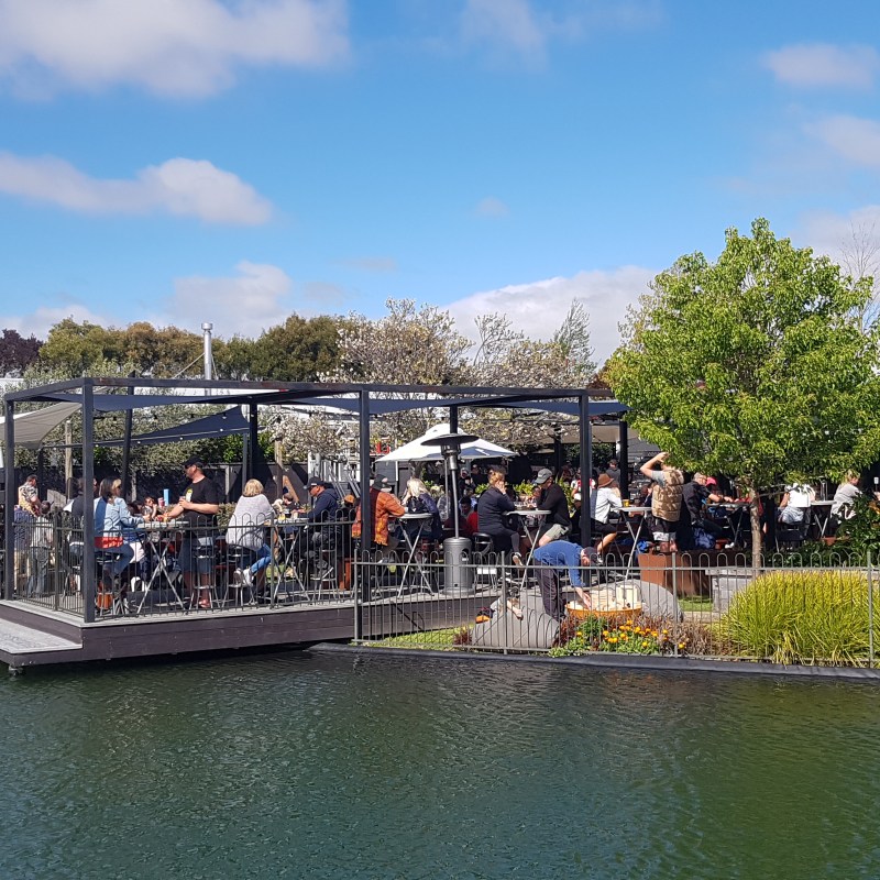 Outdoor dining area beside a pond with people seated under umbrellas and pergolas.