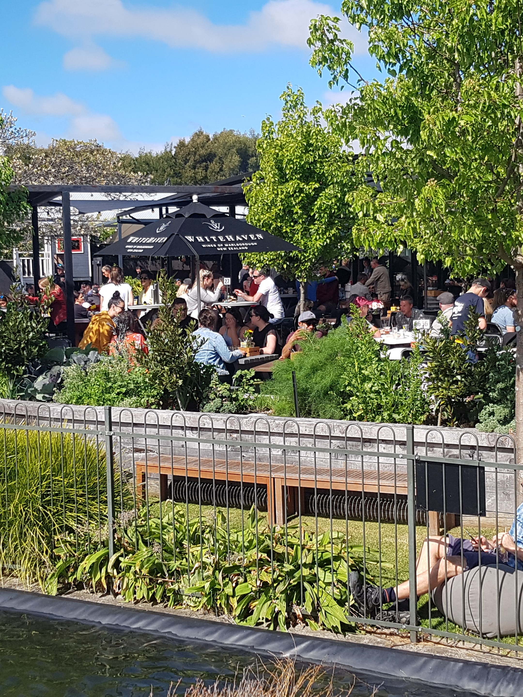 Outdoor cafe with people dining; a man relaxes on a bean bag near plants.