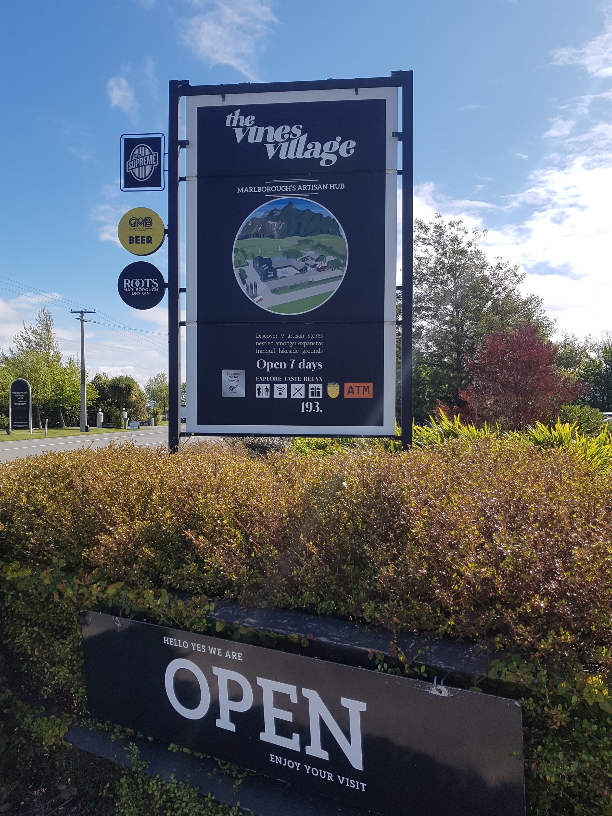 Signboard for The Vines Village with lush greenery and 'Open' sign below it.
