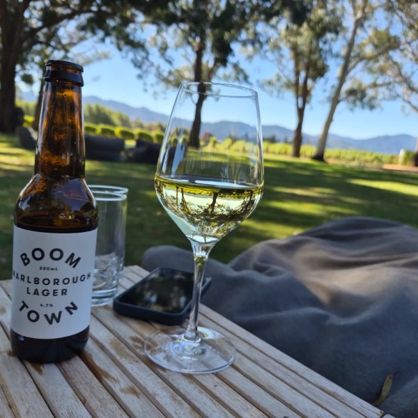 Bottle of beer and glass of wine on a wooden table outdoors with trees in the background.