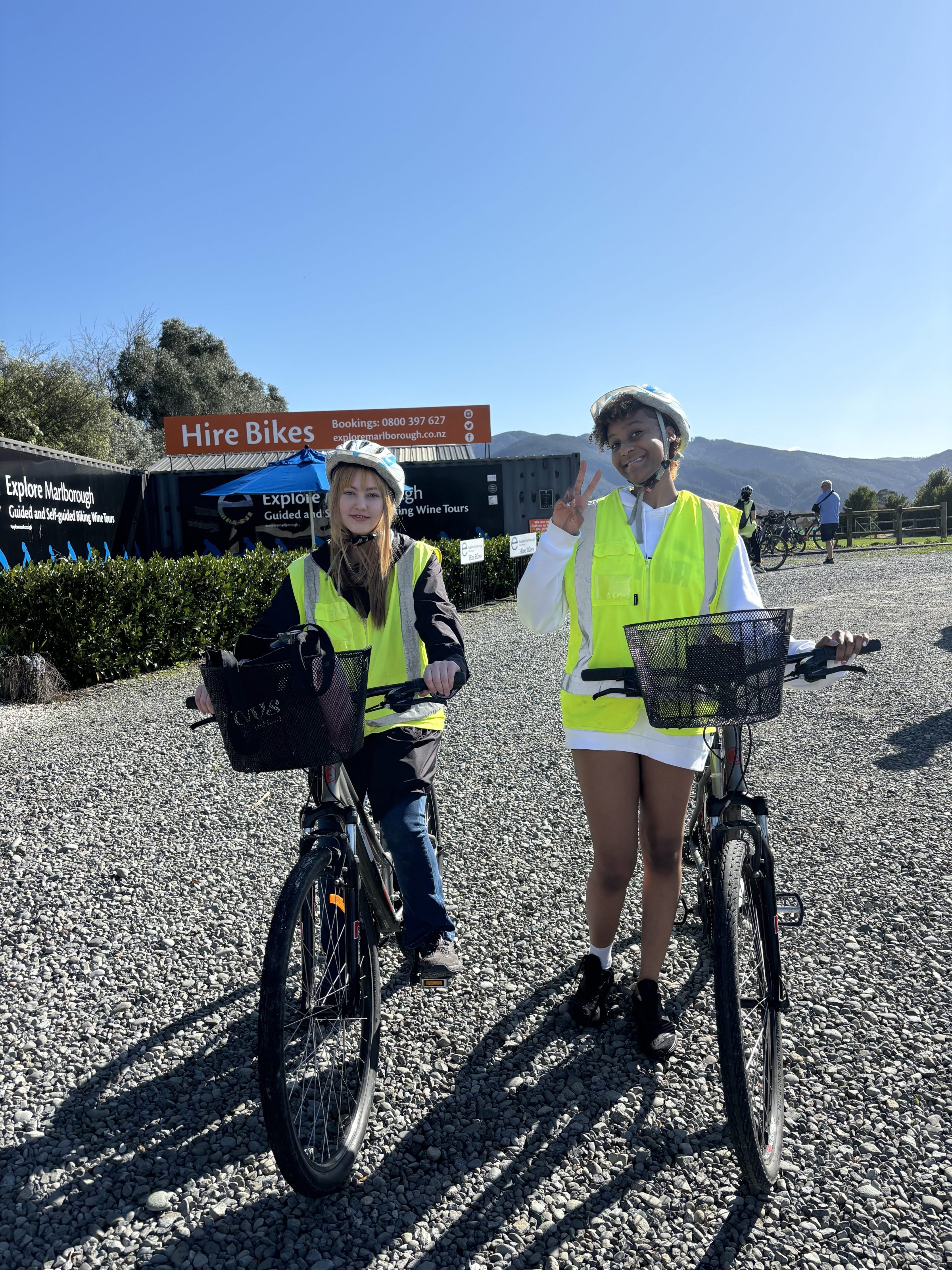 Two people in safety vests with bikes on a gravel path, mountain in background.