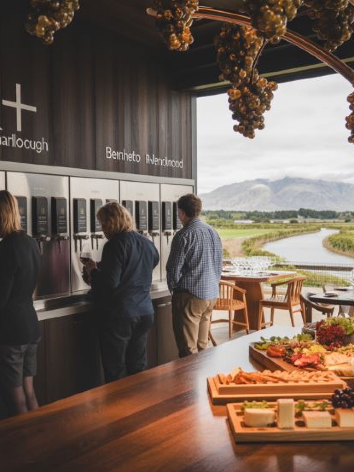 People at a wine tasting station with a landscape view and a table of appetizers.