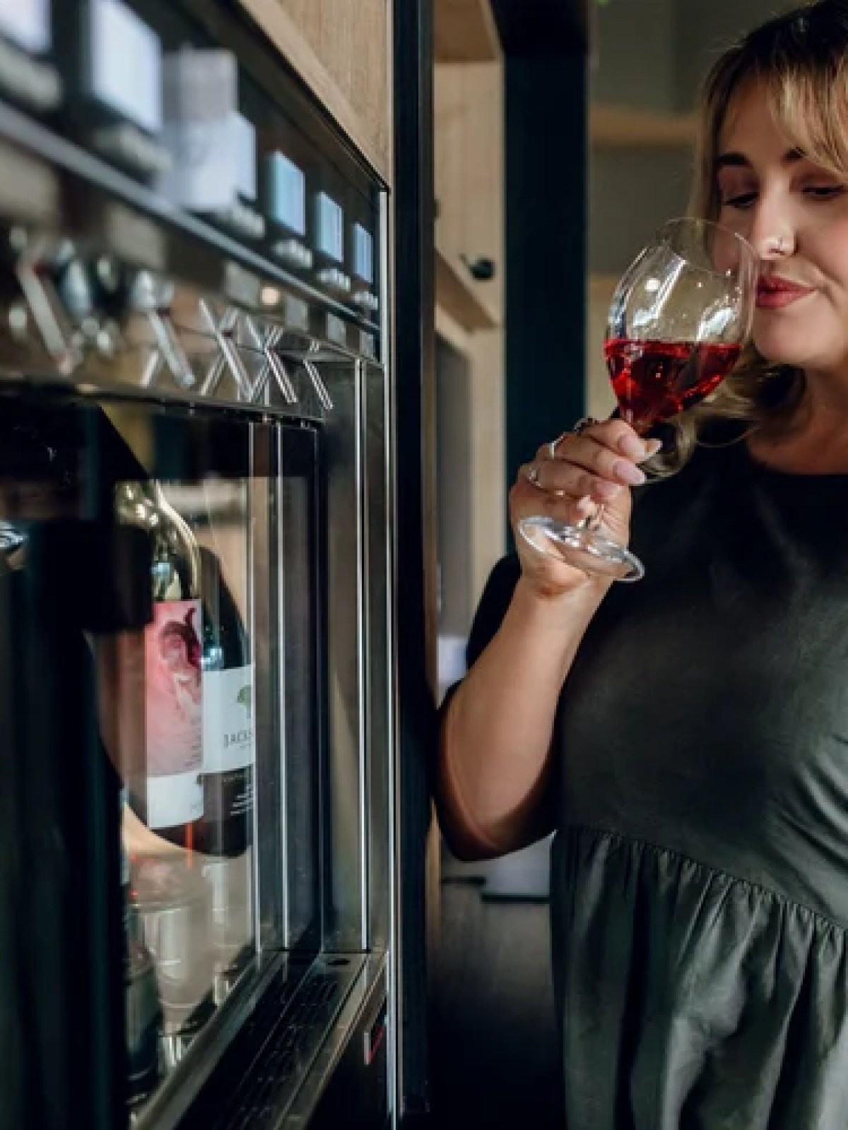 Woman in green dress smelling a glass of red wine near a wine dispenser.