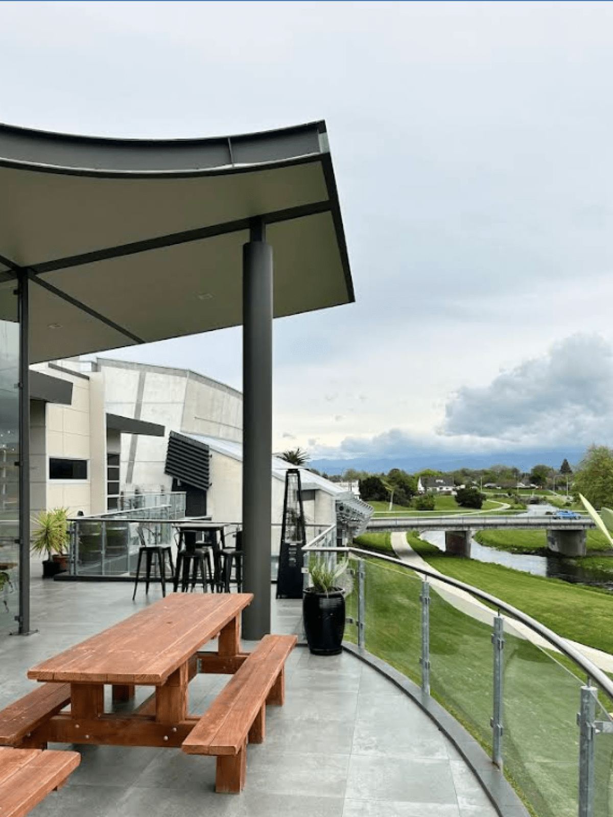 Modern patio with wooden table overlooking a river and bridge, surrounded by lush green grass and cloudy sky.