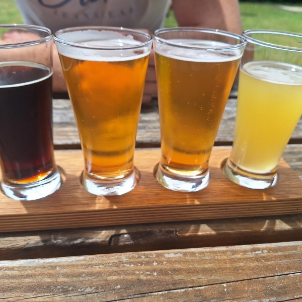 Four beer samples in glasses on a wooden tasting board on an outdoor table.