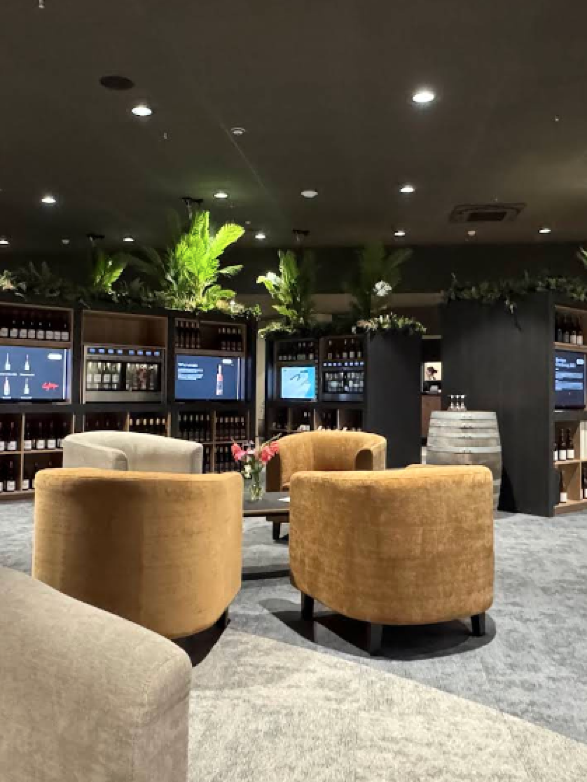 Lounge area with armchairs, plants, and wine displays under warm lighting in a spacious setting.