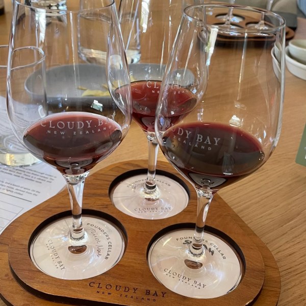 Three glasses of red wine on a wooden board labeled 'Cloudy Bay'.