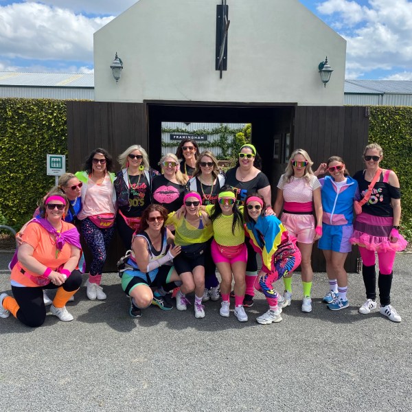 Group of people in colorful 80s-style workout outfits posing outdoors.