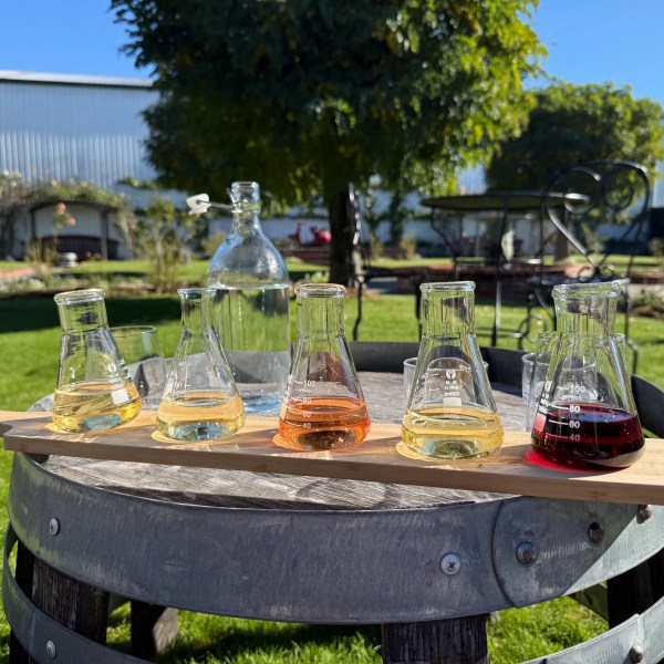 Five small beakers with colored liquids on a wooden tray outdoors on a barrel table.