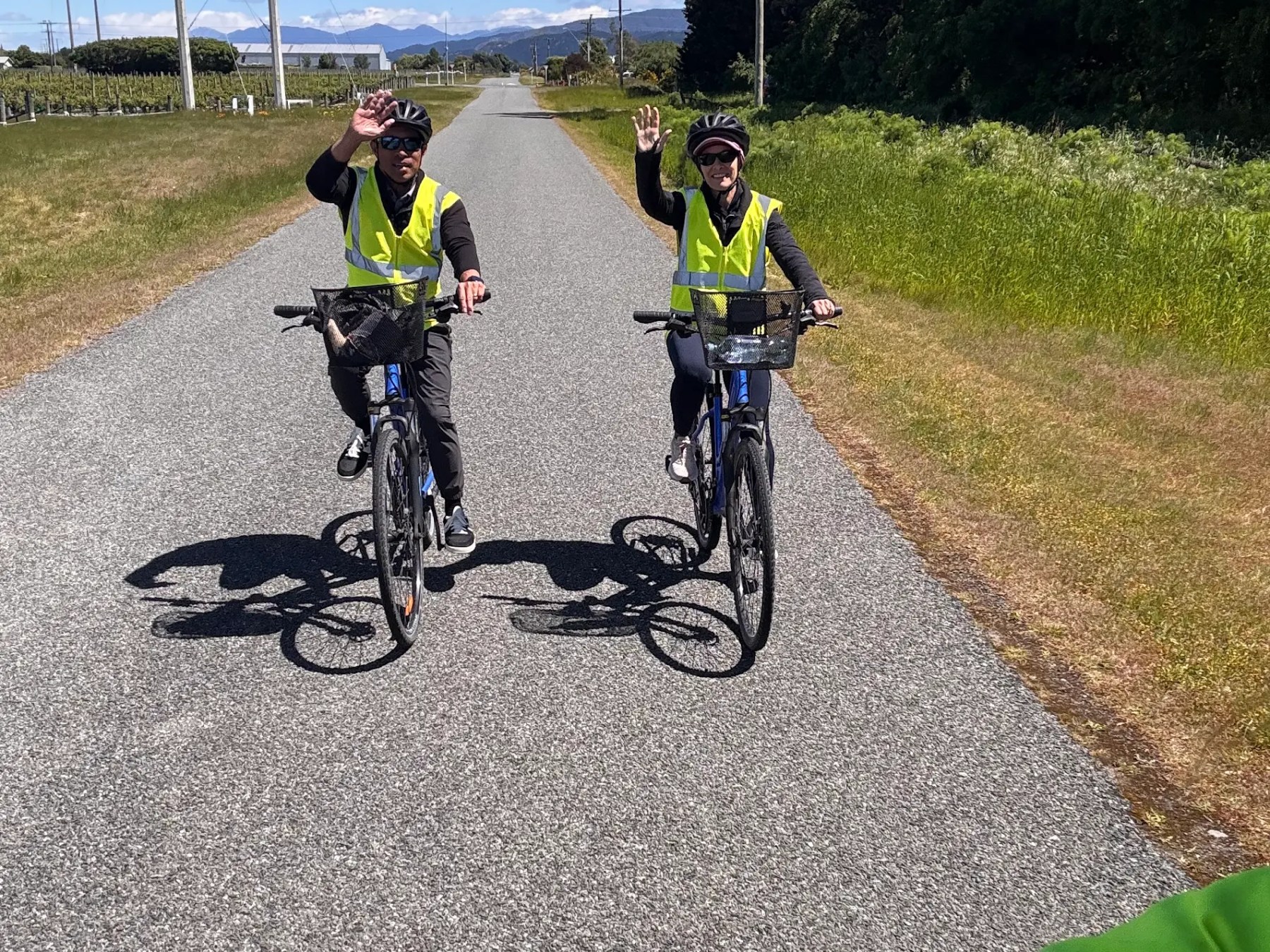 Two cyclists in yellow vests waving on a rural road under a clear blue sky.
