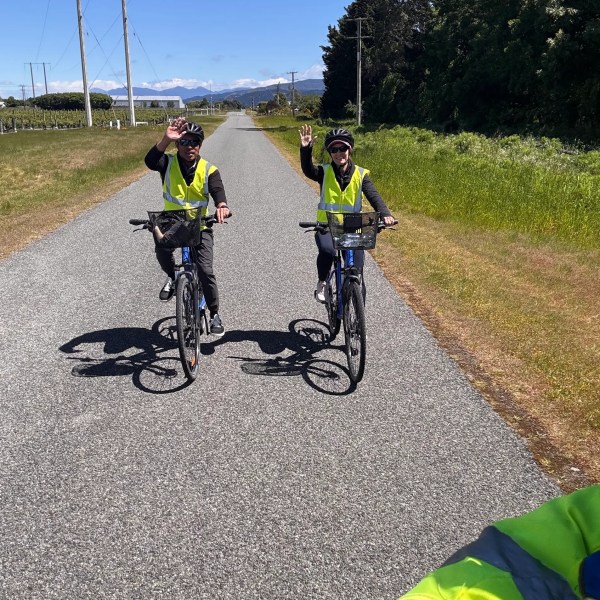 Two cyclists in yellow vests waving on a rural road under a clear blue sky.