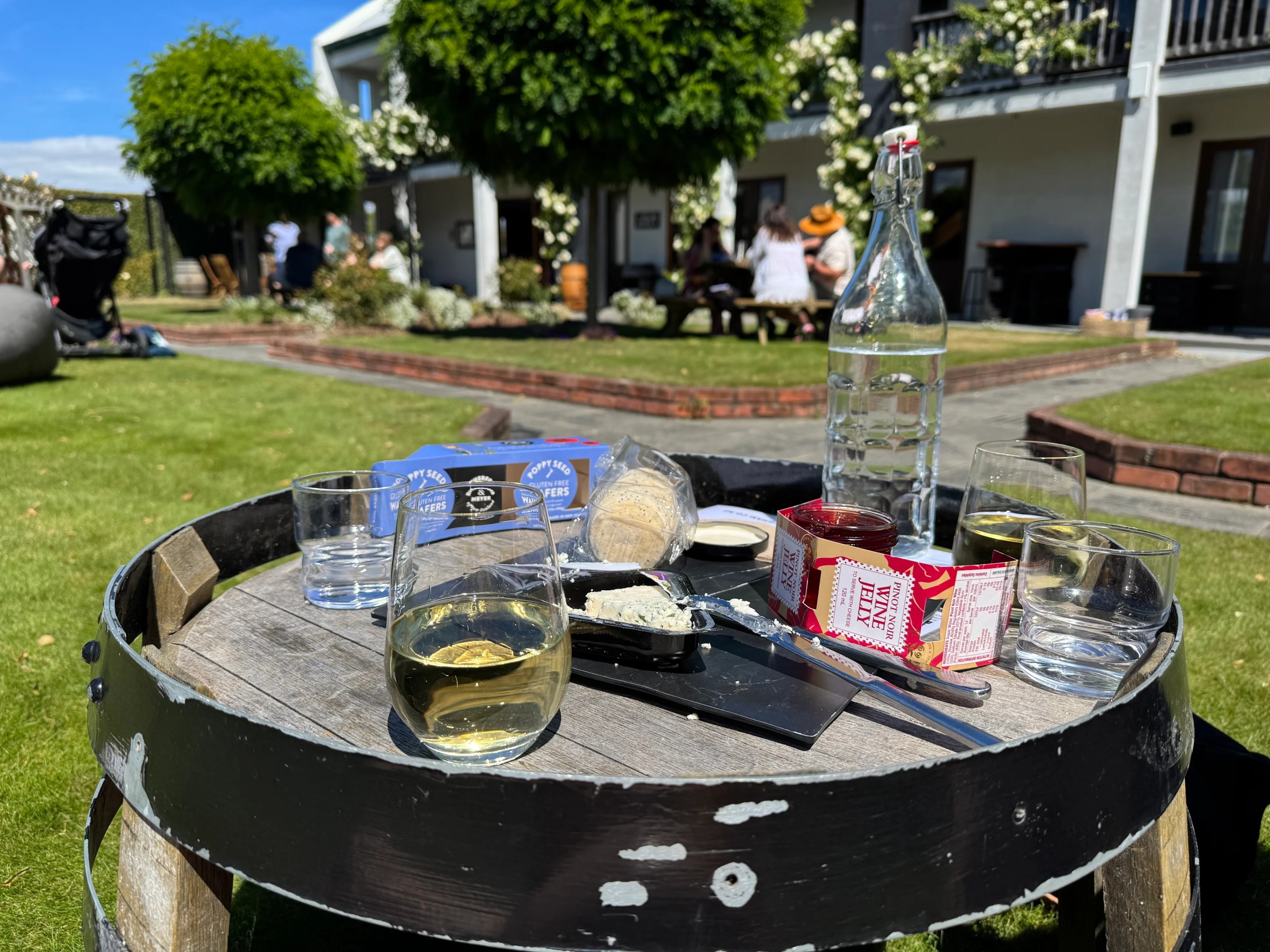 Outdoor table with wine, cheese, crackers, and wine jelly on a sunny day, people in background.