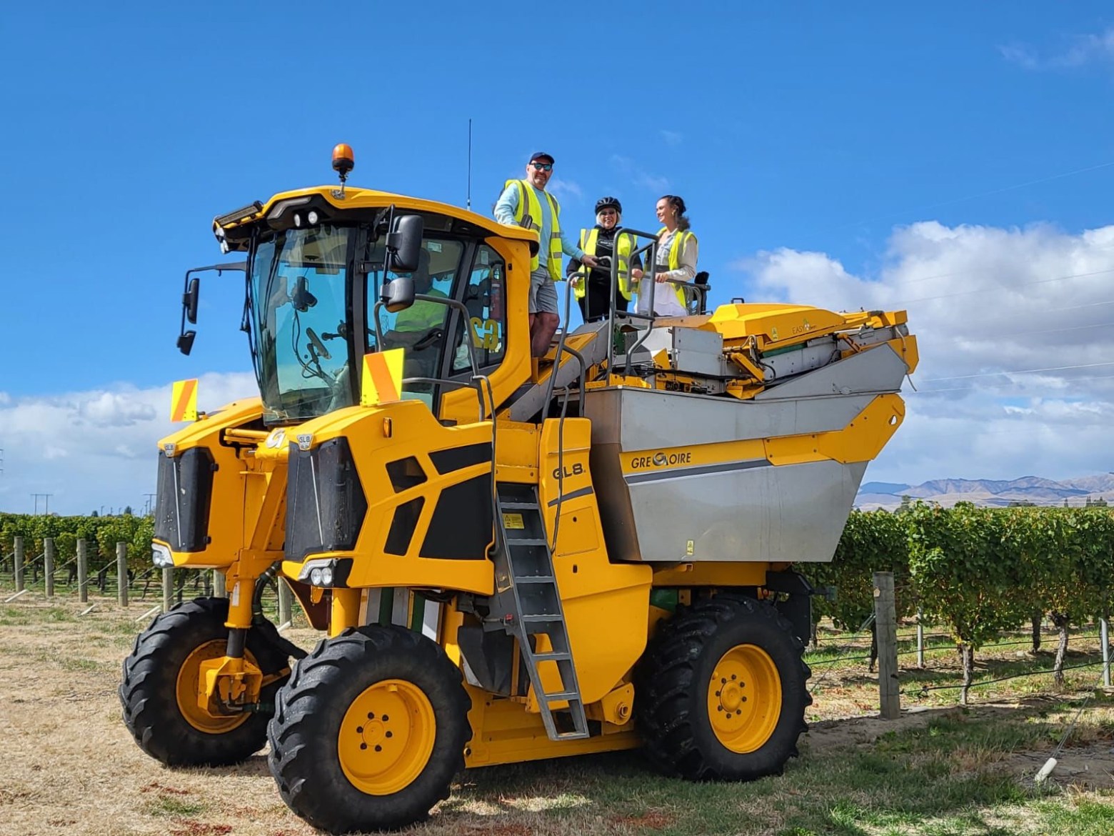 Four people on a large yellow agricultural machine in a vineyard.