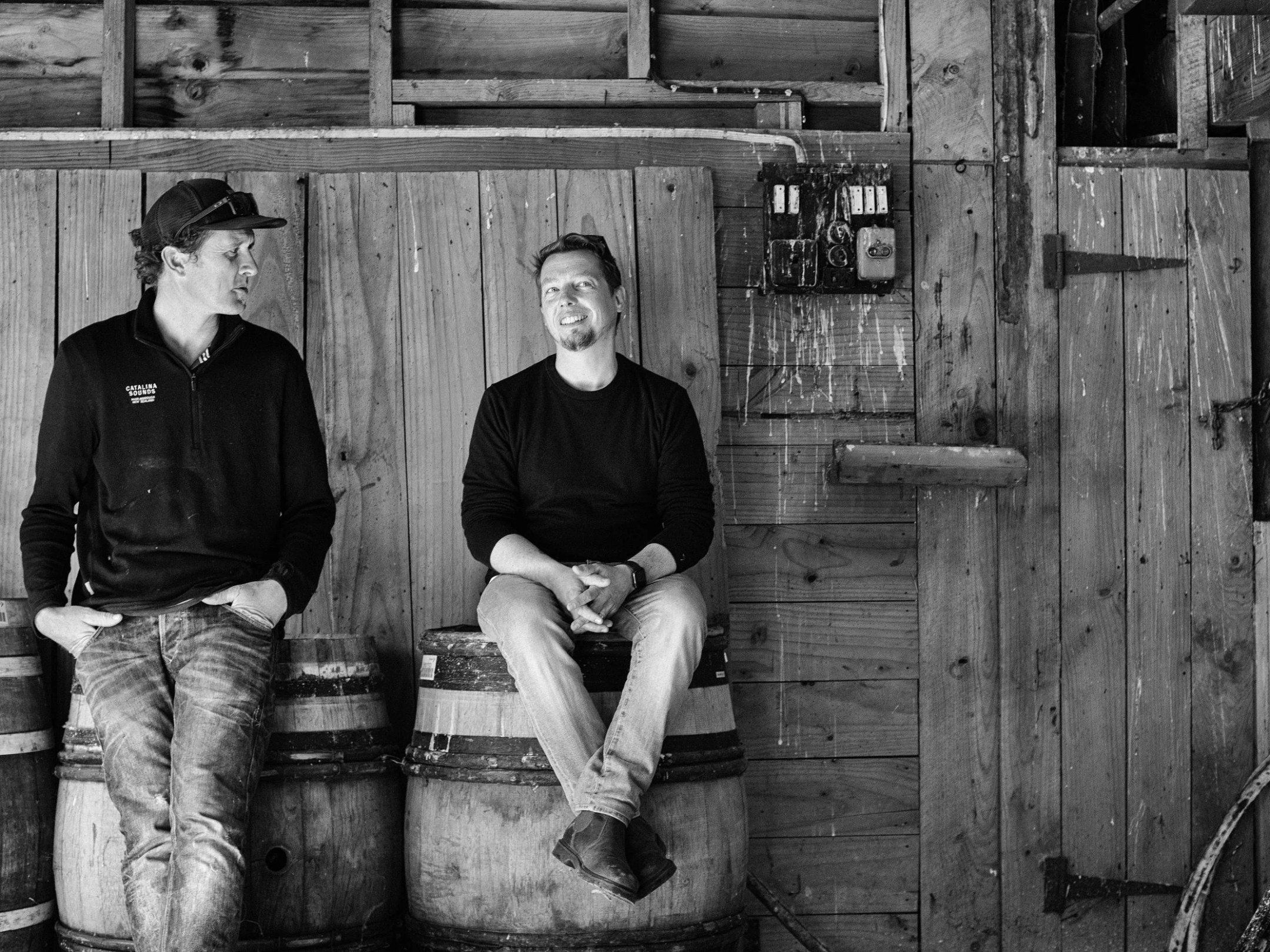 Two people sitting on barrels in a wooden barn, smiling and talking.