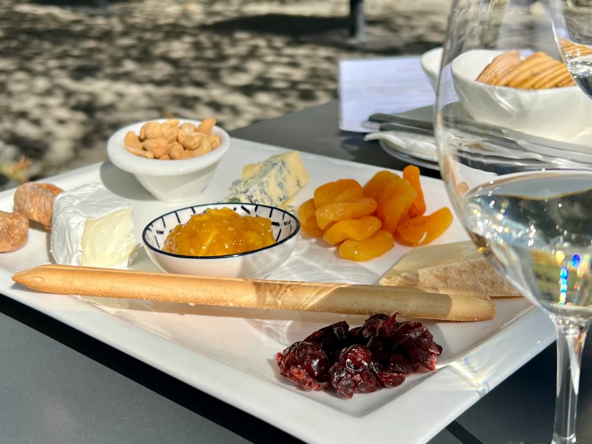 Cheese and fruit platter with wine glass on a sunny outdoor table.