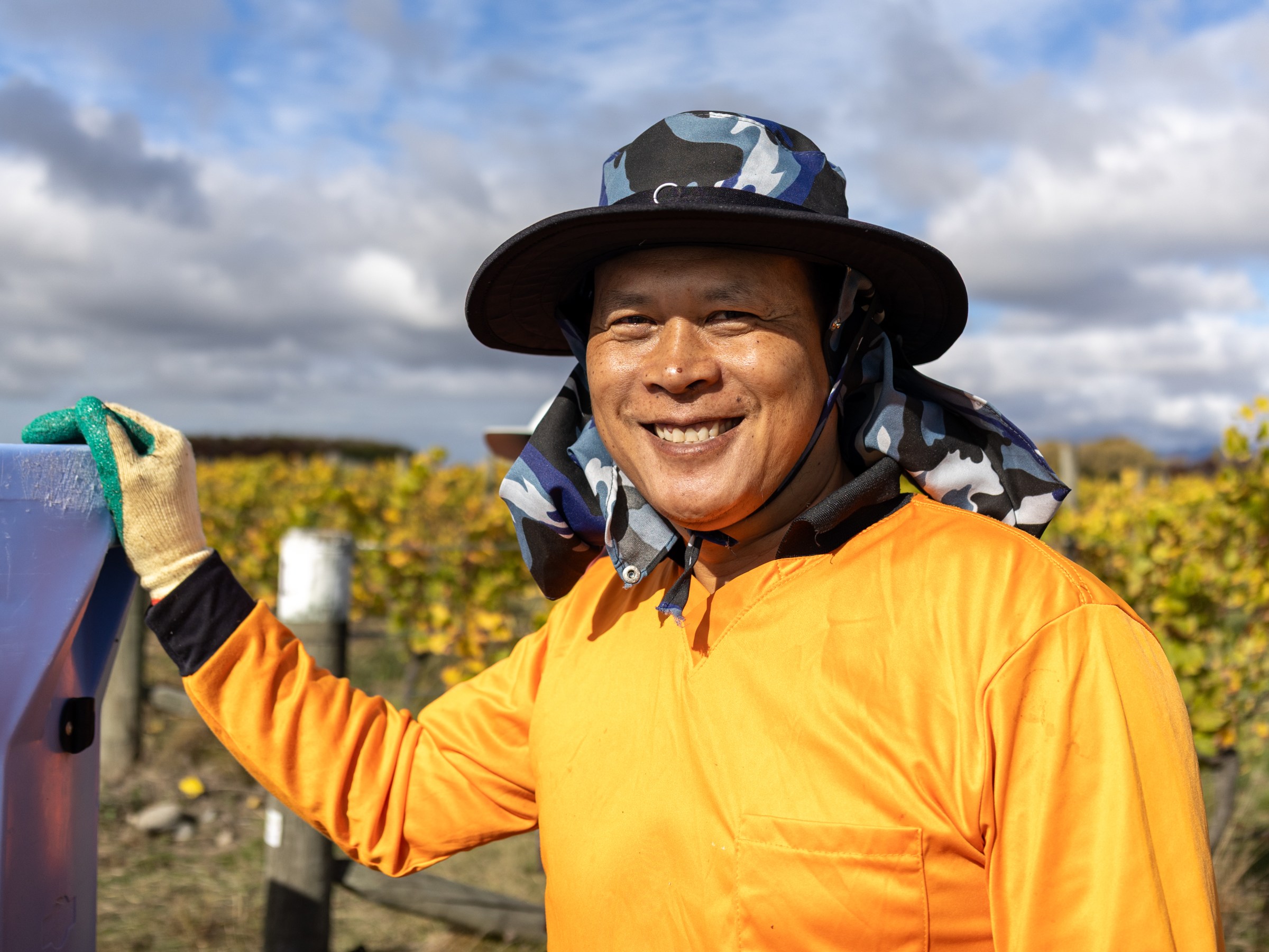Person in orange shirt and hat smiles in a vineyard under cloudy skies.