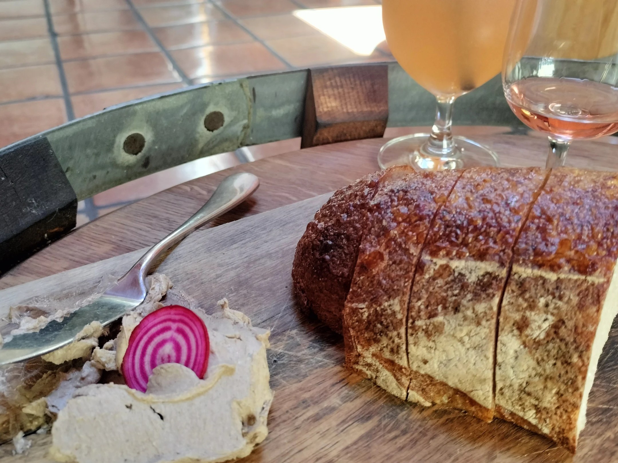 Sliced bread, a glass of beverage, and pâté with a slice of beet on a wooden board.