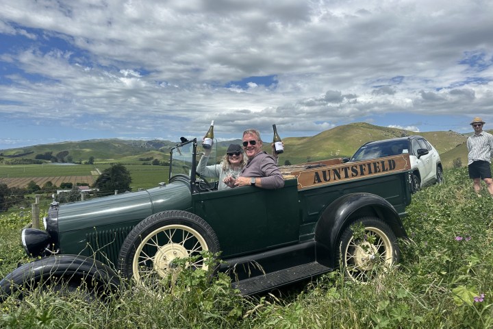 People in a vintage car on a grassy hill, holding wine bottles, with cloudy sky and hills in the background.