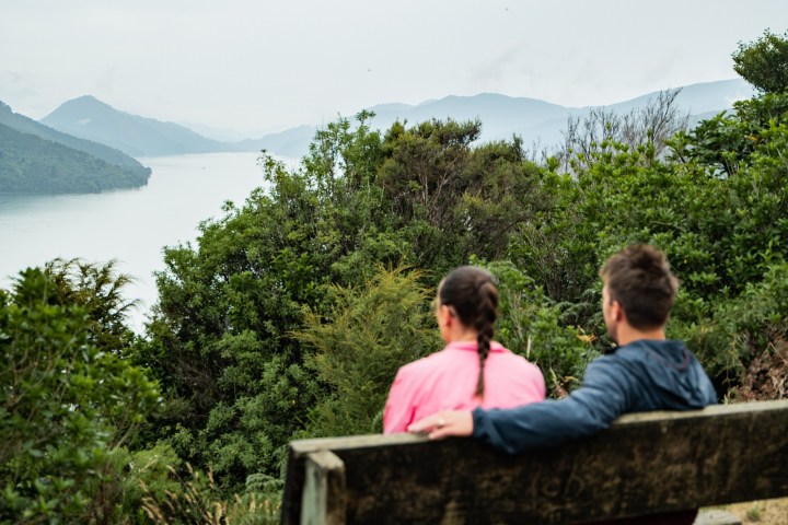 Couple sitting on a bench overlooking a scenic lake and mountains.