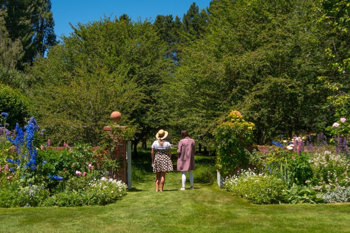 Two people walking in a lush garden toward trees under a blue sky.